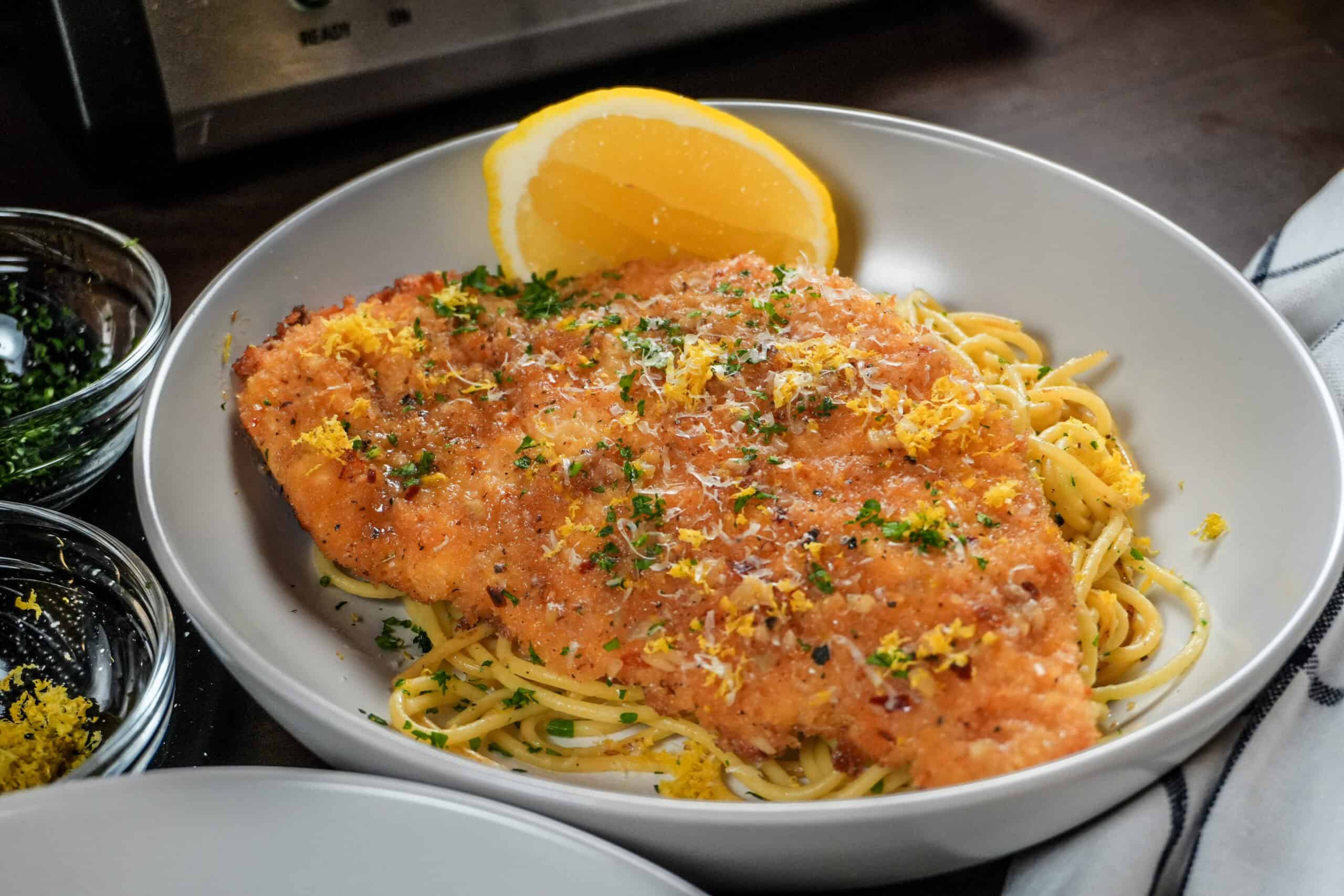 Delicious baked chicken cutlet atop garlic olive oil pasta with fresh herbs and lemon wedge.